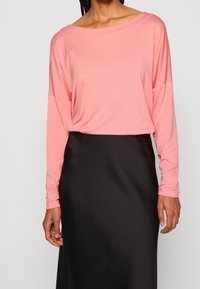 Langärmliges, locker sitzendes Oberteil aus weichem, rosa Stoff, kombiniert mit einem glatten schwarzen Rock. Das Shirt hat einen weiten Ausschnitt und abgesenkte Schultern.