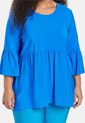 Vrouw draagt een felblauwe losse top met een geplooide taille en uitlopende ellebooglange mouwen, gecombineerd met een bijpassende blauwe broek.
