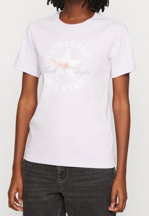 Personne portant un t-shirt Converse All Star violet clair avec un logo étoile fané et un jean noir, debout devant un fond uni.