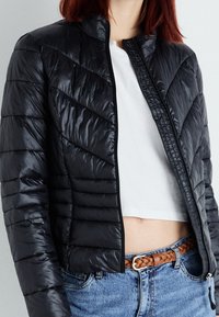 Zwarte gewatteerde jas met een verticale en chevron gestikte design. Heeft een hoge kraag en ritsluiting, gedragen over een witte crop top en blauwe jeans.