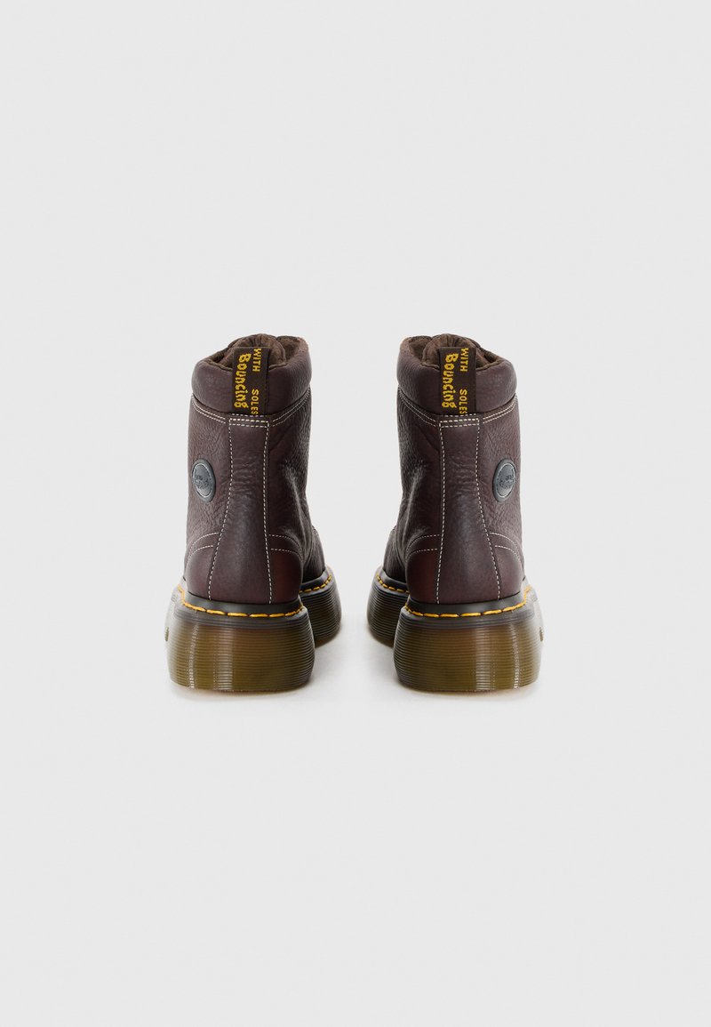 Bottines en cuir marron avec un col rembourré, bout rond, coutures jaunes et une semelle translucide robuste. Présente un logo sur le côté.