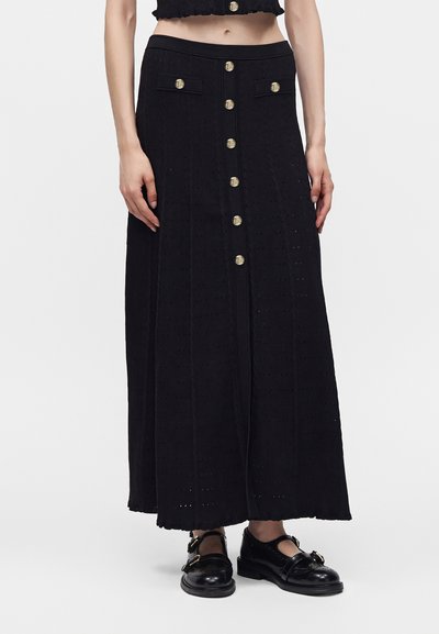 Jupe longue noire taille haute avec boutons dorés à l'avant et poches à rabat, portée avec des chaussures Mary Jane noires sur une personne debout.