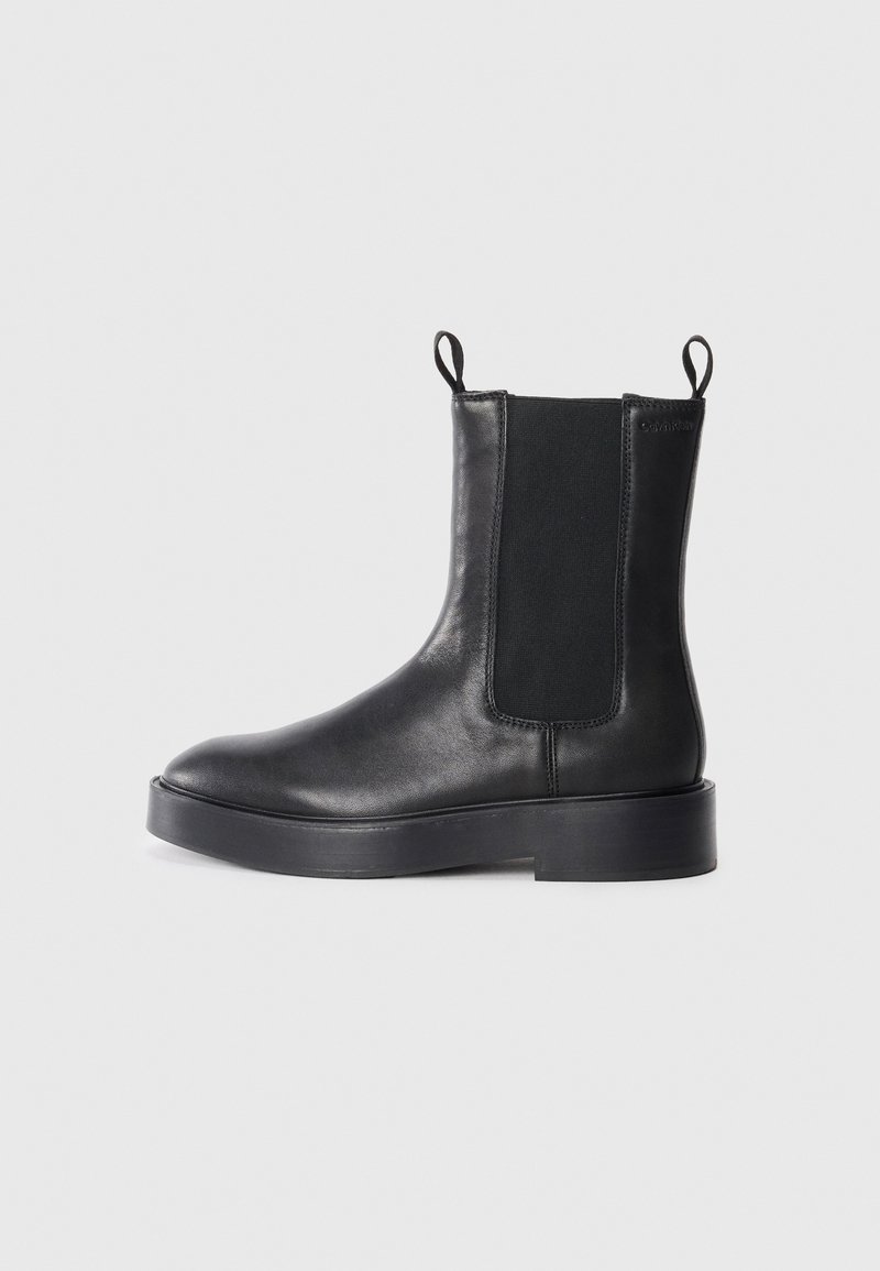 Botte Chelsea en cuir noir avec panneaux latéraux élastiques, languettes de traction en haut et semelle épaisse, présentée de profil sur un fond gris clair.