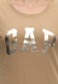 Кафява риза с металически сребърни букви, изписващи GAP на гърдите, видими от врата до горната част на торса.
