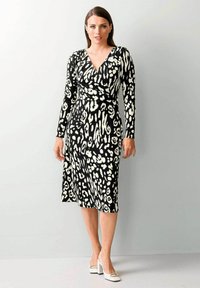 Schwarz-weißes Wickelkleid mit langen Ärmeln, das ein Leopardenmuster aufweist, knielang geschnitten und körperbetont ist. Weiße Schuhe mit Blockabsatz.