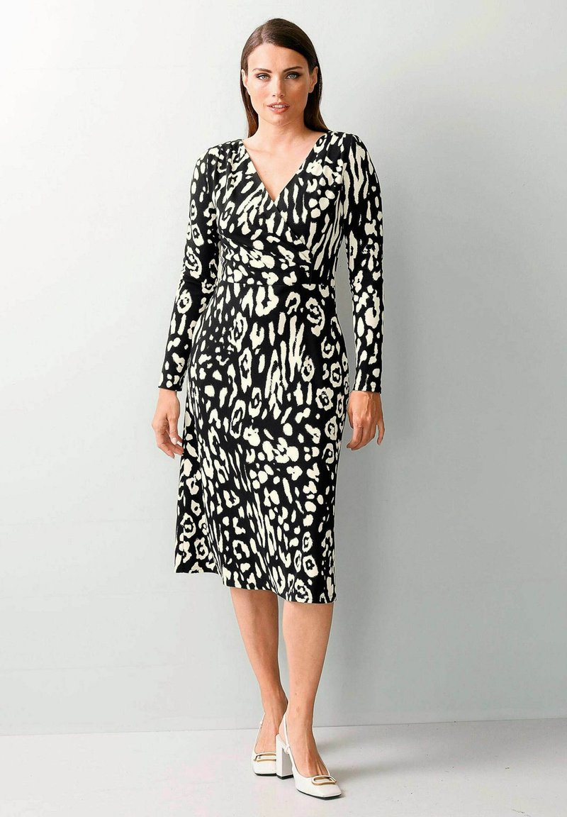 Schwarz-weißes Wickelkleid mit langen Ärmeln, das ein Leopardenmuster aufweist, knielang geschnitten und körperbetont ist. Weiße Schuhe mit Blockabsatz.
