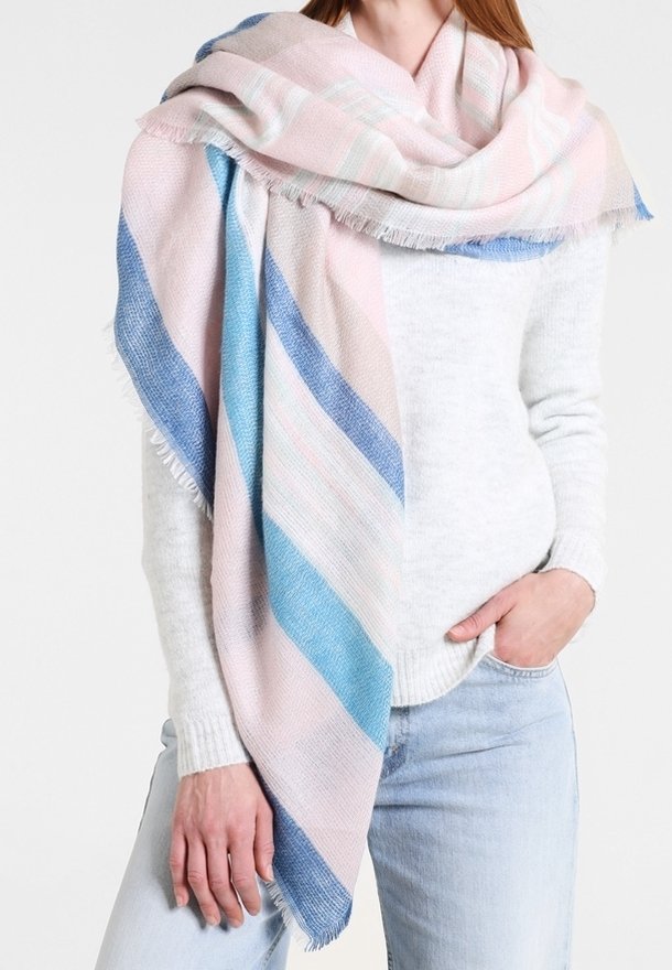 Femme portant une grande écharpe pastel rayée rose, bleu et blanc sur un pull blanc et un jean bleu clair, main dans la poche.