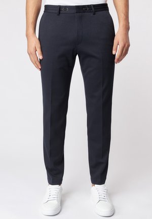 Mand iført mørkeblå slim-fit bukser kombineret med hvide sneakers, stående mod en neutral baggrund.
