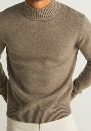 Uomo che indossa un maglione girocollo in maglia a coste color talpa con polsini e orlo a coste, abbinato a pantaloni chiari, con le mani appoggiate sui fianchi.
