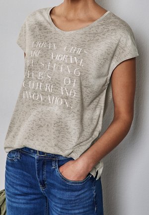Persona vistiendo una camiseta beige claro con patrón y texto sobre ciudades urbanas, combinada con vaqueros azules, mano en el bolsillo, de pie frente a una pared lisa.