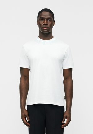 Νεαρός άντρας με σκουρόχρωμο δέρμα που φοράει απλό λευκό κοντομάνικο μπλουζάκι και μαύρο παντελόνι, στέκεται μπροστά σε απλό φωτεινό φόντο.