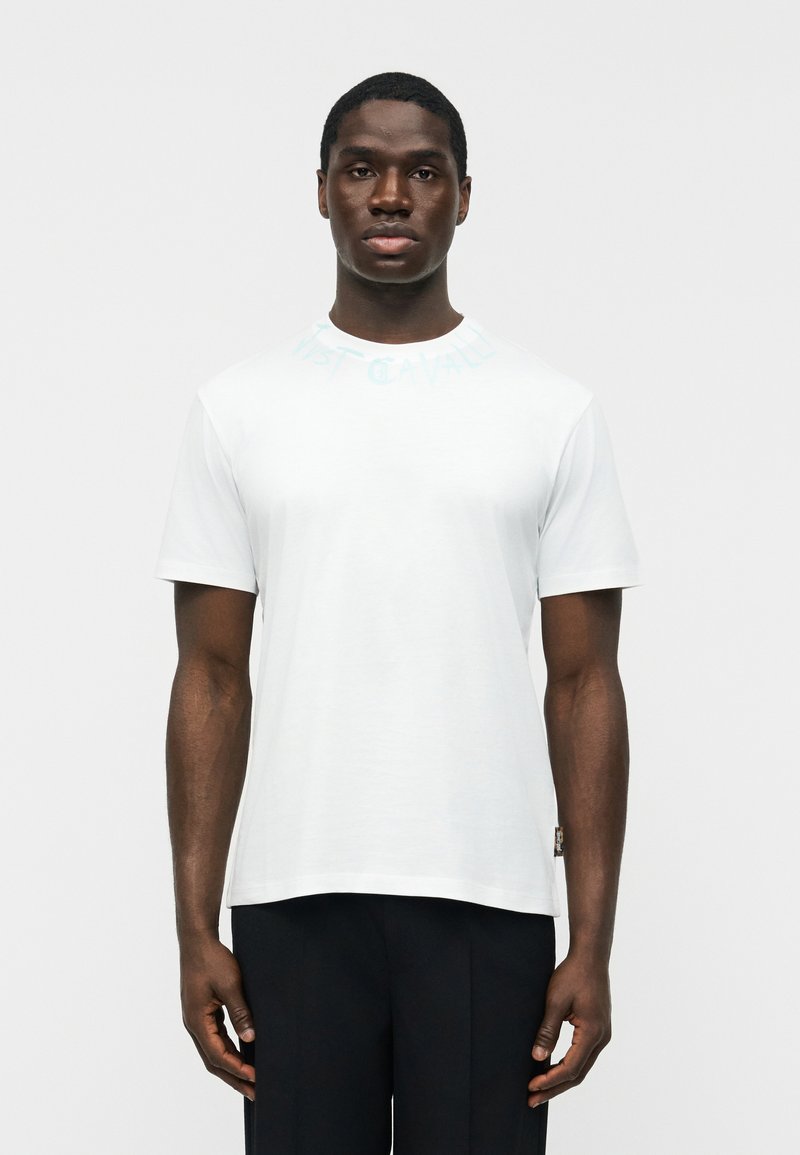 Νεαρός άντρας με σκουρόχρωμο δέρμα που φοράει απλό λευκό κοντομάνικο μπλουζάκι και μαύρο παντελόνι, στέκεται μπροστά σε απλό φωτεινό φόντο.