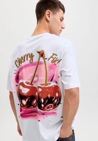 Νεαρός άνδρας που φοράει λευκό μπλουζάκι με μεγάλο γραφικό δύο λαμπερών κερασιών και το κείμενο "Cherry Fix!" στην πλάτη.