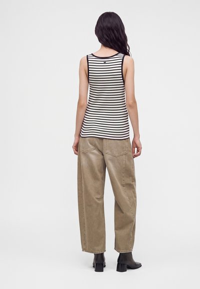Femme aux longs cheveux foncés portant un haut sans manches rayé noir et blanc, un pantalon beige ample et des bottes noires à talons, debout, de dos.