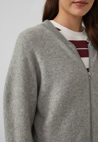 Grauer Wollcardigan mit Reißverschluss, geripptem Kragen und weicher Textur. Darunter ist ein weißes Shirt mit burgunderfarbenen Streifen teilweise sichtbar.