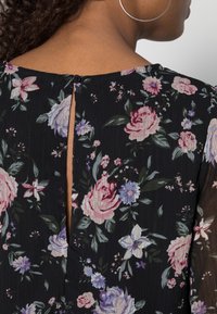 Schwarze Bluse mit floralem Muster, die große rosa Rosen, lila Blumen und grüne Blätter zeigt. Mit einem Tropfenausschnitt hinten und einem Knopfverschluss. Leichter Stoff.