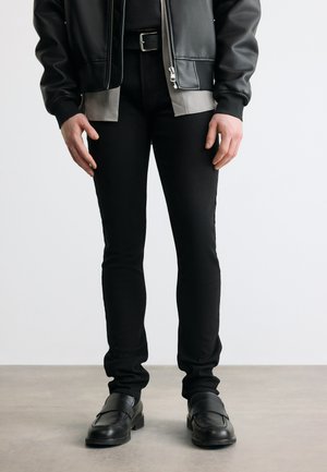 Person trägt schwarze Slim-Fit-Hose, schwarze Lederjacke, graues Hemd, schwarzen Gürtel und schwarze Lederschuhe und steht auf einem grauen Boden.