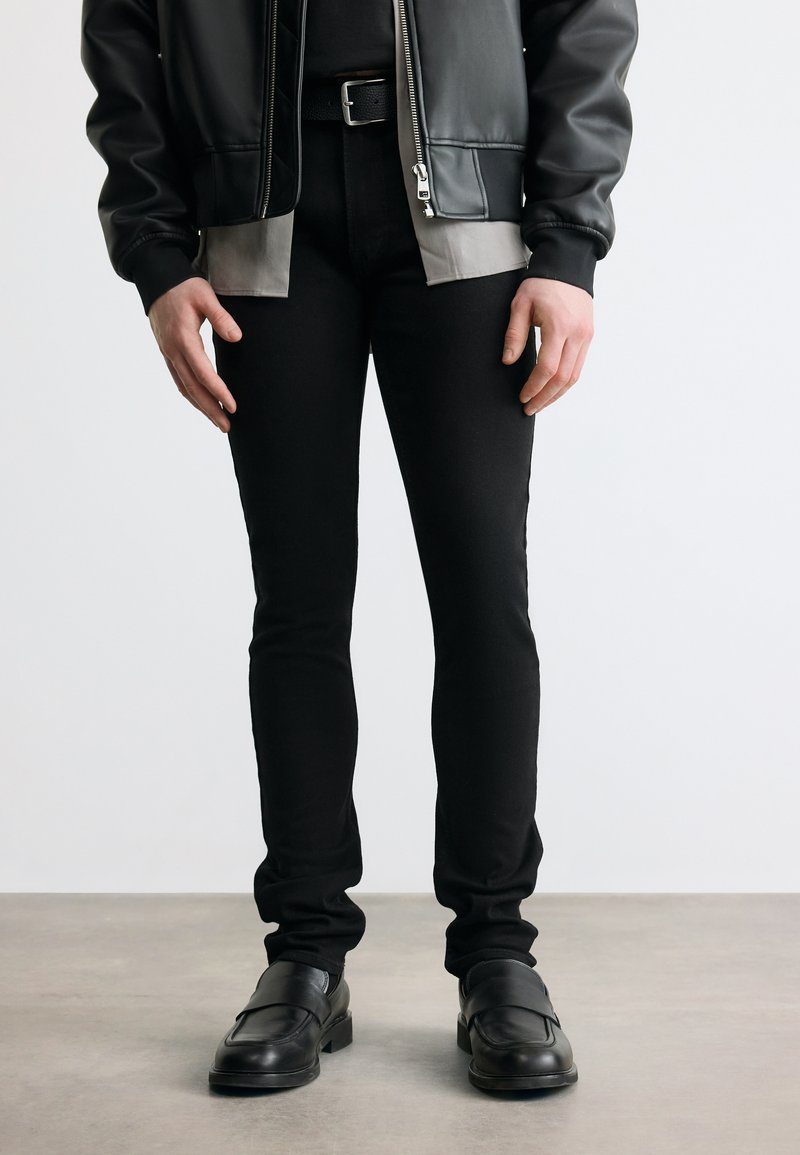 Άτομο που φοράει μαύρο στενό παντελόνι, μαύρο δερμάτινο μπουφάν, γκρι πουκάμισο, μαύρη ζώνη και μαύρα δερμάτινα παπούτσια, στέκεται σε γκρι δάπεδο.