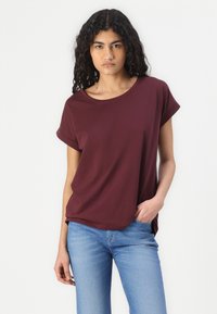 Tricou scurt, mărimea maron, cu guler rotund, croială relaxată și mâneci rulate, purtat împreună cu blugi ușor albaștri. Textura țesăturii este netedă.
