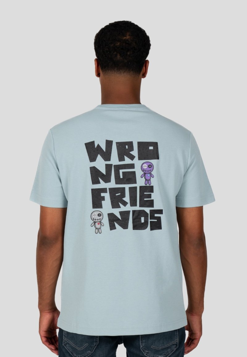 Vaaleansininen puuvillapaidan, jossa on suuri musta teksti "VÄÄRÄT YSTÄVÄT" ja kaksi värikästä sarjakuvamaista nukke-grafiikkaa selkäpuolella.