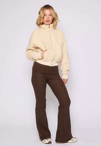 Giacca bomber in pile beige con chiusura a zip e polsini a costine, abbinata a pantaloni marrone scuro a zampa. La modella posa su uno sfondo neutro.