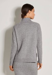 Frau mit dunklem Haar in einem tiefen Dutt, die einen hellgrauen gestrickten Rollkragenpullover und ein passendes Rock trägt, steht vor einer schlichten hellen Wand.