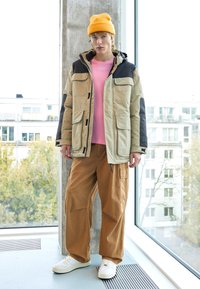 Lett og flerlommet beige og svart vinterjakke, rosa skjorte under, lysebrune vide bukser og hvite joggesko med en oransje beanie.