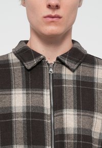 Plaid zip-up jacket featuring a dark brown and beige color scheme, flannel material, and a pointed collar with a metallic zipper.