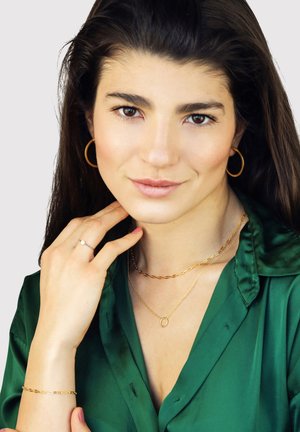 Young woman with long dark hair wears gold hoop earrings, layered gold necklaces, a gold bracelet, and a green satin blouse, touching her neck.