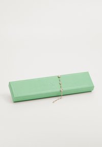 Caixa retangular verde com uma textura suave, contendo uma pulseira de ouro com várias pedras redondas transparentes.