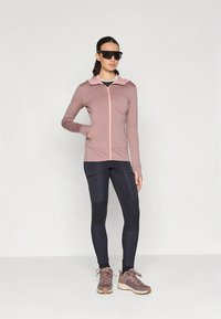 Casaco com capuz rosa claro com fecho éclair contrastante, combinado com leggings azul-marinho. A modelo usa sapatilhas desportivas castanhas e óculos de sol pretos oversize.