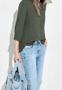 Blouse texturée vert olive à manches trois-quarts, associée à un jean en denim bleu clair, tenant un sac fourre-tout en tissu bleu clair.