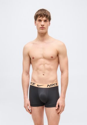 Jaunas vyras stovi be marškinėlių, dėvi juodus „Nike“ bokserius su smėlio spalvos juosmuo, žiūri tiesiai į priekį priešais paprastą foną.