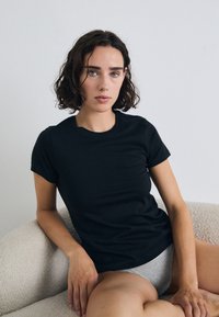 Jeune femme aux cheveux courts et bouclés assise sur un canapé texturé de couleur claire, portant un t-shirt noir et des sous-vêtements gris, regardant directement l'appareil photo.
