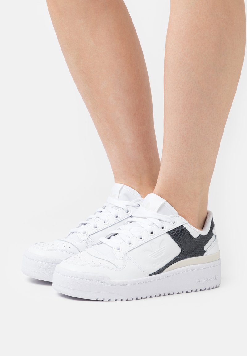 Baskets basses blanches avec des accents latéraux texturés noirs portées pieds nus sur un fond clair uni.