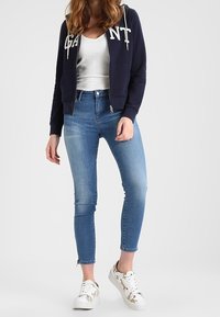 Felpa con zip blu navy con lettering bianco, top bianco, jeans skinny blu chiaro e sneakers con pattern e dettagli floreali.