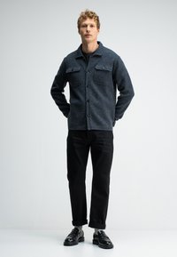 Grau gestrickte Hemdjacke mit zwei vorderen Taschen, schwarzen Knöpfen und einem Kragen, kombiniert mit schwarzen Hosen mit hochgekrempelten Säumen und schwarzen Slippern.