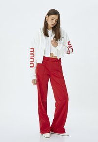 Sudadera blanca recortada con texto y acentos rojos, combinada con pantalones de pierna ancha rojos brillantes de cintura alta. Zapatillas blancas completan el conjunto.