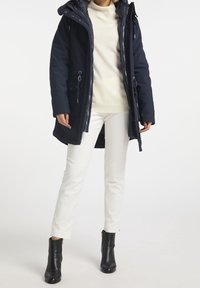 Parka azul marino con capucha removible, con cierre de cremallera y cordón, dos bolsillos laterales, llevada sobre un suéter crema y pantalones blancos.