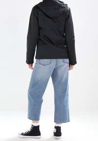 Osoba v černé kapucové bundě, světle modrých zkrácených džínách, bílých ponožkách a černých vysokých teniskách Converse, stojící zády k objektivu.