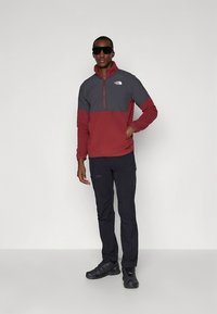 Rote und graue Fleecejacke mit Reißverschluss und hohem Kragen, getragen mit schwarzen Hosen, dunklen Sneakers und Sonnenbrille.