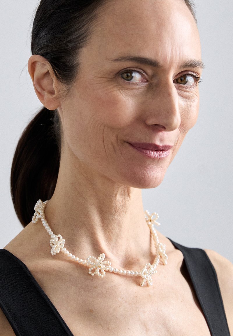 Λευκό κολιέ από μαργαρίτες με λουλουδένια αξεσουάρ, που αποτελείται από πλεγμένο νήμα με μικρούς μαργαρίτες, εκτεθειμένο σε μοντέλο με ουδέτερο φόντο.