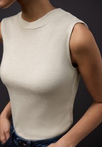 Krémový top bez rukávů s vysokým límcem, vyrobený z texturované látky s jemným leskem. Kombinován s tmavě modrými džínami.