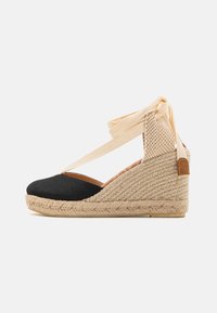 Espadrilles a zeppa con tela nera, suola in juta naturale intrecciata in contrasto, con cinturini crema incrociati e un accento decorativo marrone chiaro sul tallone.