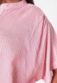 Růžová a bílá pruhovaná blůza s vysokým límcem, krátkými rukávy a jemnou texturou látky, která obsahuje delikátní vertikální šití.