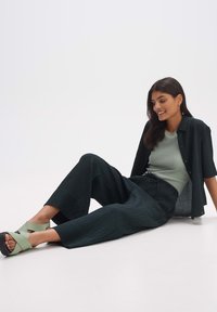 Donkergroene, gestructureerde broek met wijde pijpen, gecombineerd met een lichtgroene, nauwsluitende top en een zwart overhemd met korte mouwen met knopen. Groene sandalen.