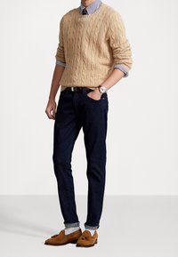 Beige kabelstickad tröja, blå och vit randig skjorta under, mörka jeans, bruna mocka loafers. Klocka på handleden, händerna i fickorna.