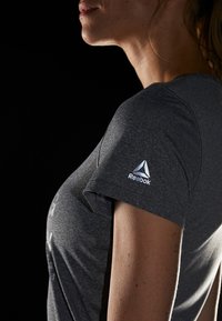Camiseta atlética gris con cuello redondo, mangas cortas y un logo reflectante de Reebok en la manga izquierda. Textura de tela suave.