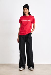 Červené bavlnené tričko s logom "Calvin Klein Jeans", skombinované s čiernymi širokými nohavicami a čiernym čiapkou. Obsahuje strieborné tenisky.