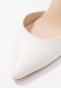 Chaussure blanche à bout pointu en cuir lisse avec un design découpé sur les côtés et un intérieur beige, montrant un style épuré et minimaliste.